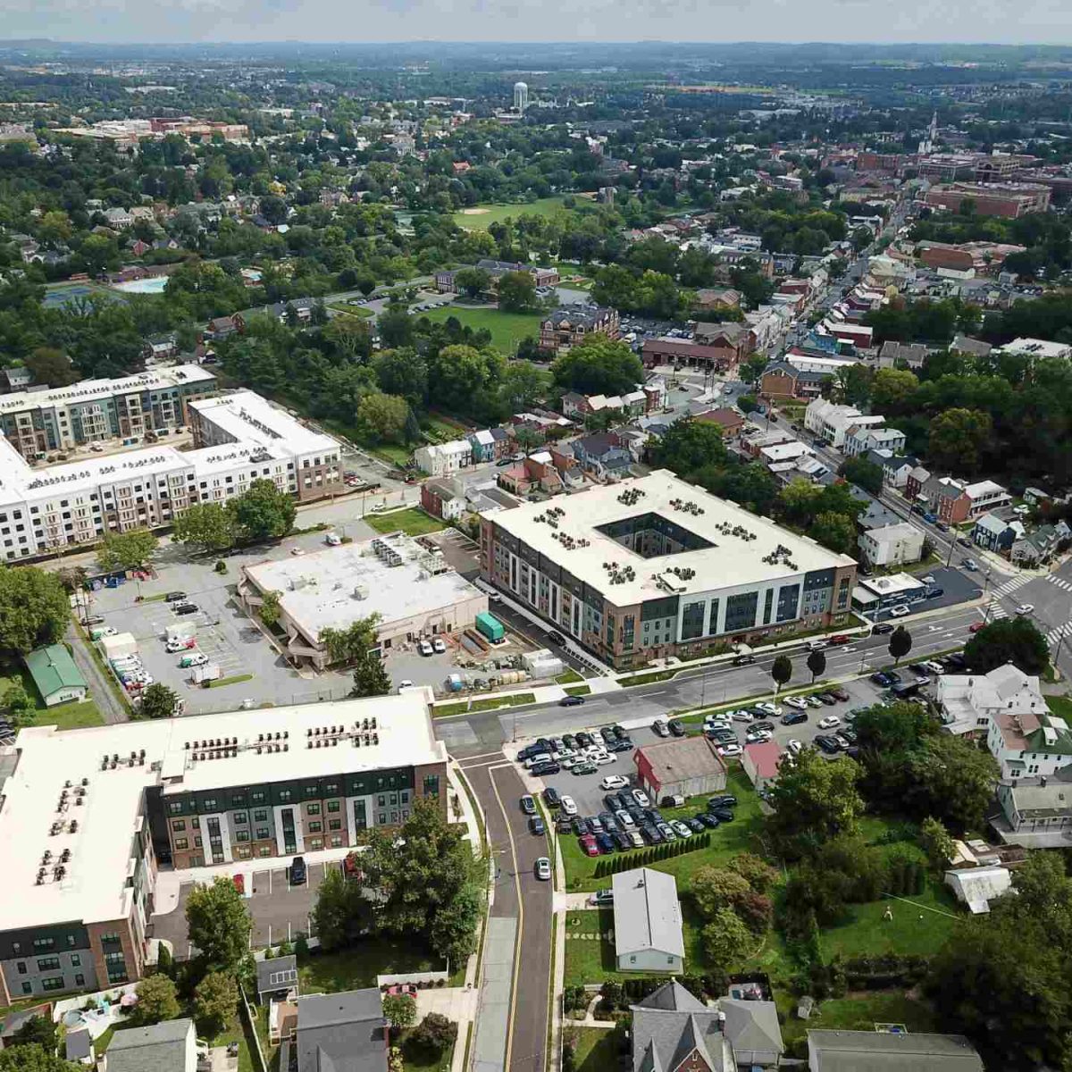 Aerial view of new apartment buildings with white rooftops surrounded by a wooded residential town with streets and homes.
