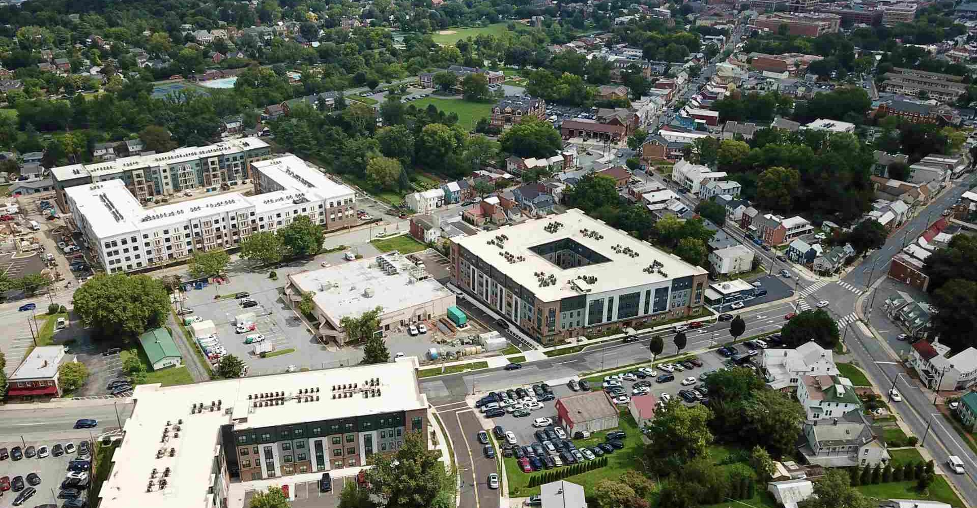 Aerial view of new apartment buildings with white rooftops surrounded by a wooded residential town with streets and homes.